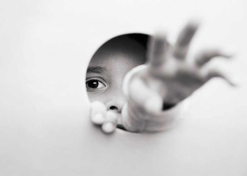 a child reaching through a hole in a wall, stretching out an arm and making eye contact with the camera