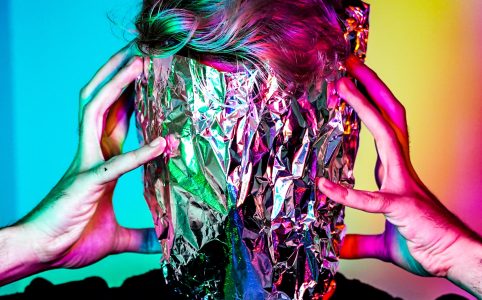 a person with aluminum foil obscuring their face, with hands on both sides of their face, in multicolored light