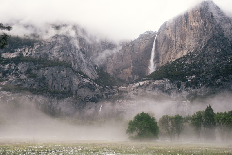 a closeup of a mountain range, partially covered in fog. some trees can be seen on the ground below.