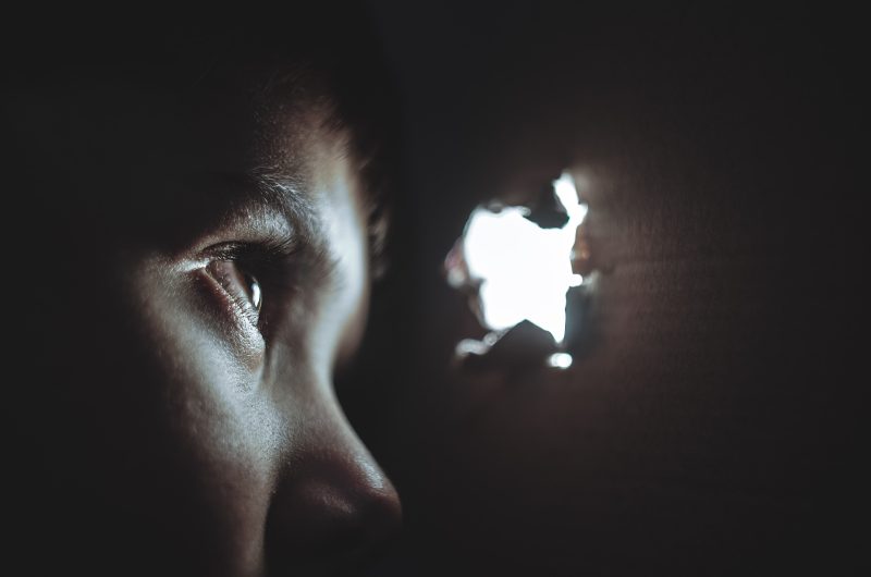 a child looks through a small hole from within a cardboard box. light comes through the hole in the box.