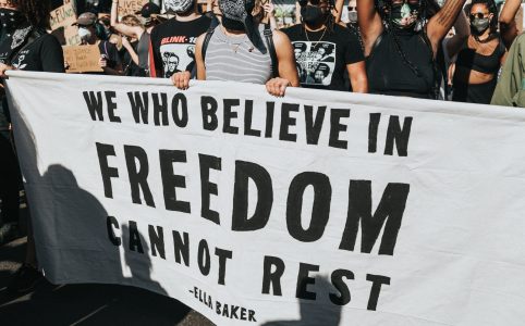 people marching with many signs, including a large sign held by several people, white with black letters that reads "we who believe in freedom cannot rest" by Ella Baker