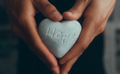 two brown hands holding a heart-shaped stone carved with the word 'Hope'