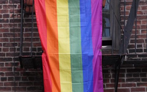 a rainbow flag hangs from the metal balcony of a brick building