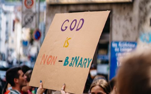 in a crowd at a peaceful protest gathering, a person in a white face mask and dark hair with glasses holds a sign that reads 'GOD IS NON-BINARY'