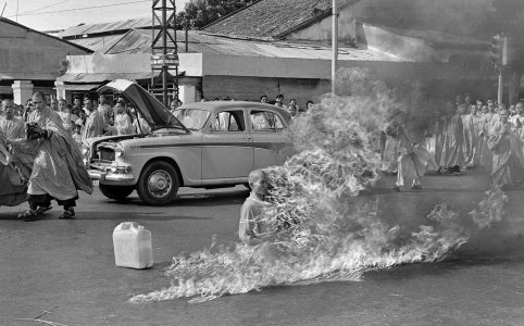 a black and white photo of a street with people in robes and a car in front of a cluster of buildings. the focal point is a monk who is actively on fire. this is the famous photo of Quảng Đức during his self-immolation