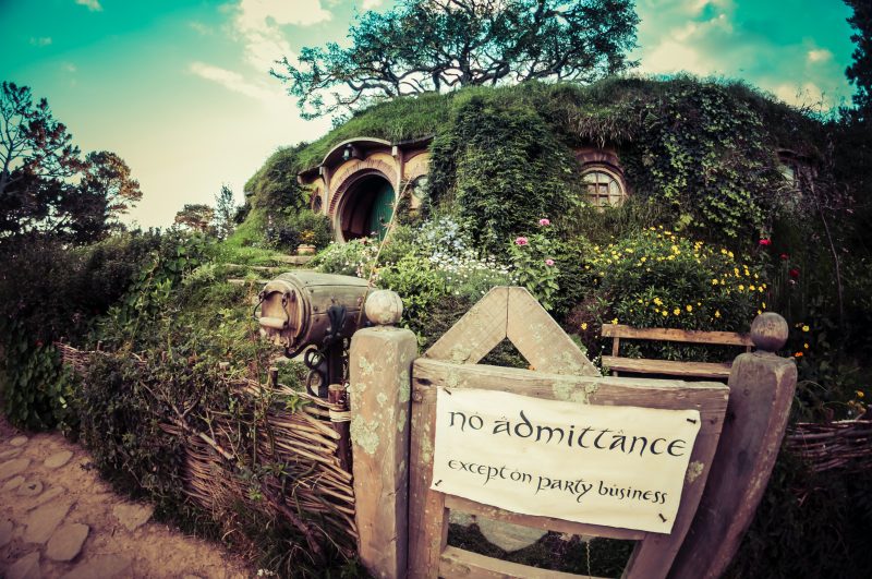 a scene of a hobbit house surrounded by flowering plants and a wicker fence, with a sign reading 'no admittance except on party business' nailed to the gate