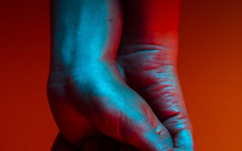 closeup of two people holding hands, palm outward, washed in blue light against a red background