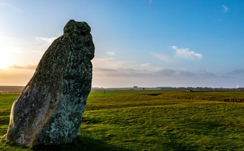 a photo of sunlight off-camera to the left, with a leaning ancient stone in front of the light. there is a grassy field and the sky is blue with some clouds.