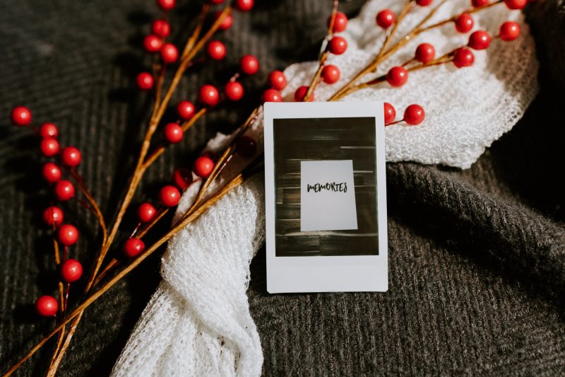 a photo of the word 'memories', framed in white, lying with a spray of red berries and a white cloth on a dark knit blanket