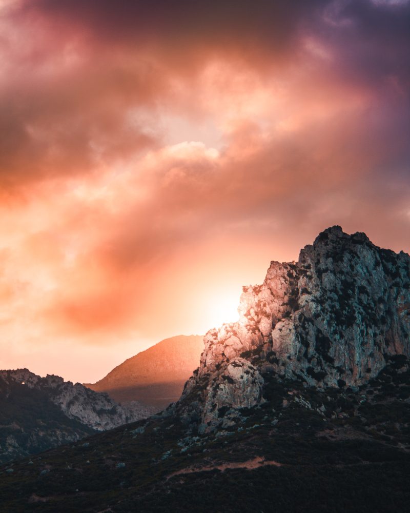 a mountain range with some snow-capped peaks. there are sun-suffused clouds behind them, and the sun glows on the lower right.