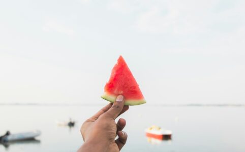 a brown-skinned hand holding a watermelon slice against a backdrop of water