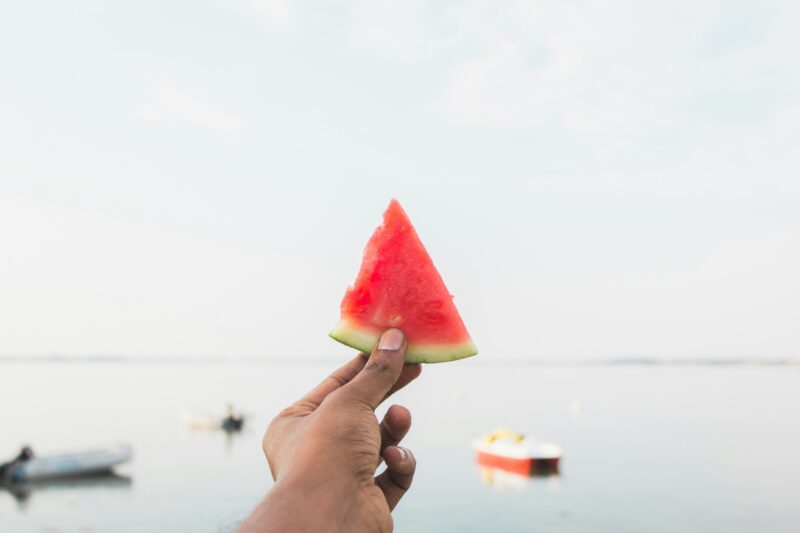 a brown-skinned hand holding a watermelon slice against a backdrop of water