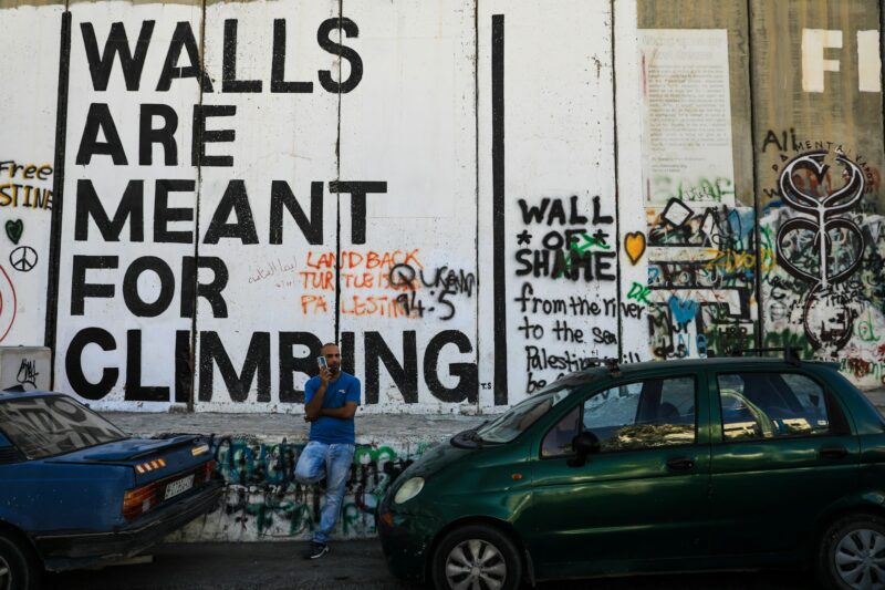 large lettering on a concrete wall: WALLS ARE MEANT FOR CLIMBING. LAND BACK TURTLE ISLAND PALESTINE. WALL OF SHAME. from the river to the sea Palestine will be free.